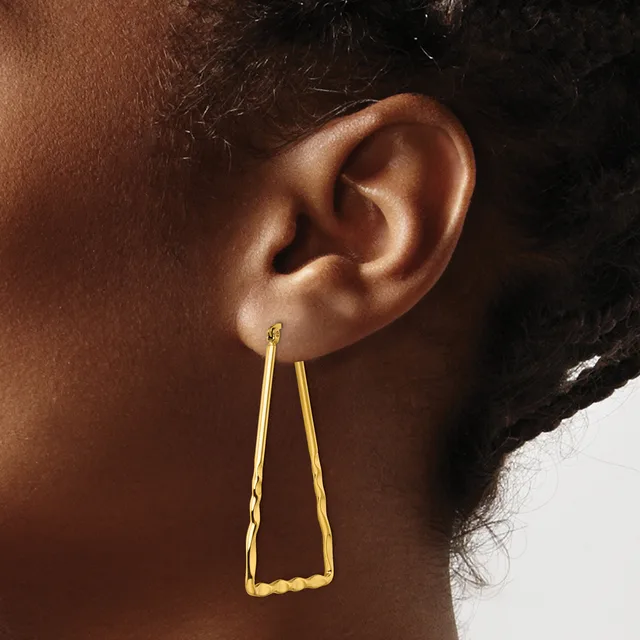 Gold trapezoid-shaped drop earrings with a textured surface worn on a person's ear against a neutral background.