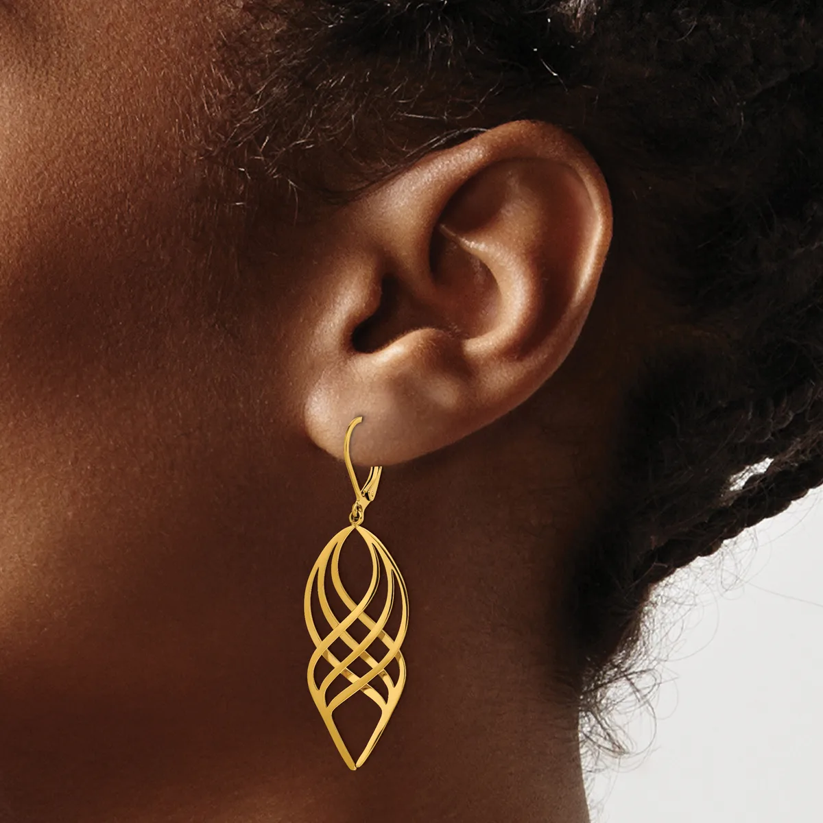 A gold-toned elongated leaf-shaped earring with interlocking geometric cutouts hangs from a wire hook on a model's ear.