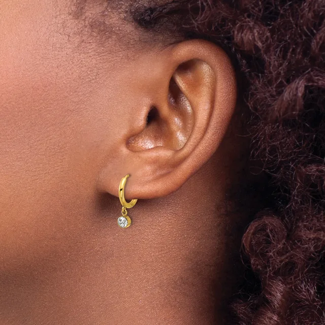 A gold hoop earring with a small round diamond charm hangs from the earlobe of a person with dark curly hair.