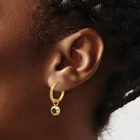 A gold hoop earring with a small circular charm hanging from it, worn on the ear of a person with dark skin tone and black hair.