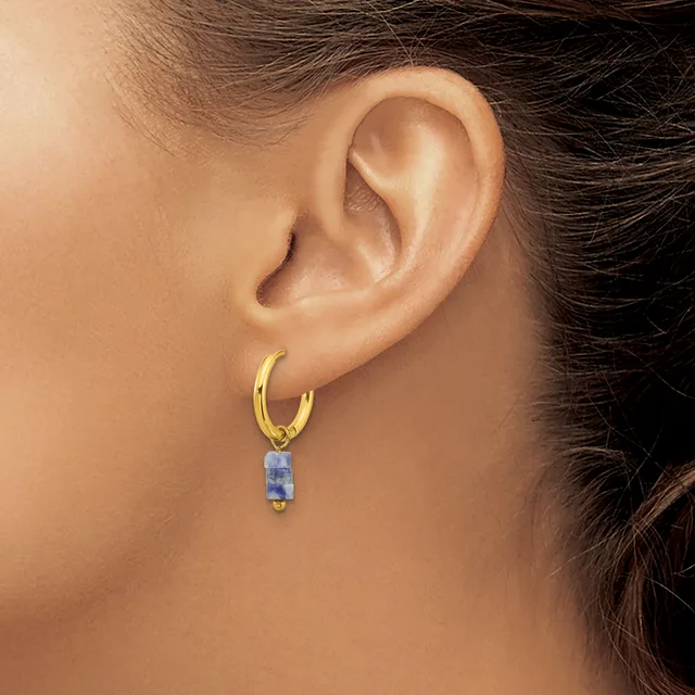 A gold hoop earring with a small rectangular blue stone pendant hangs from a woman's earlobe.