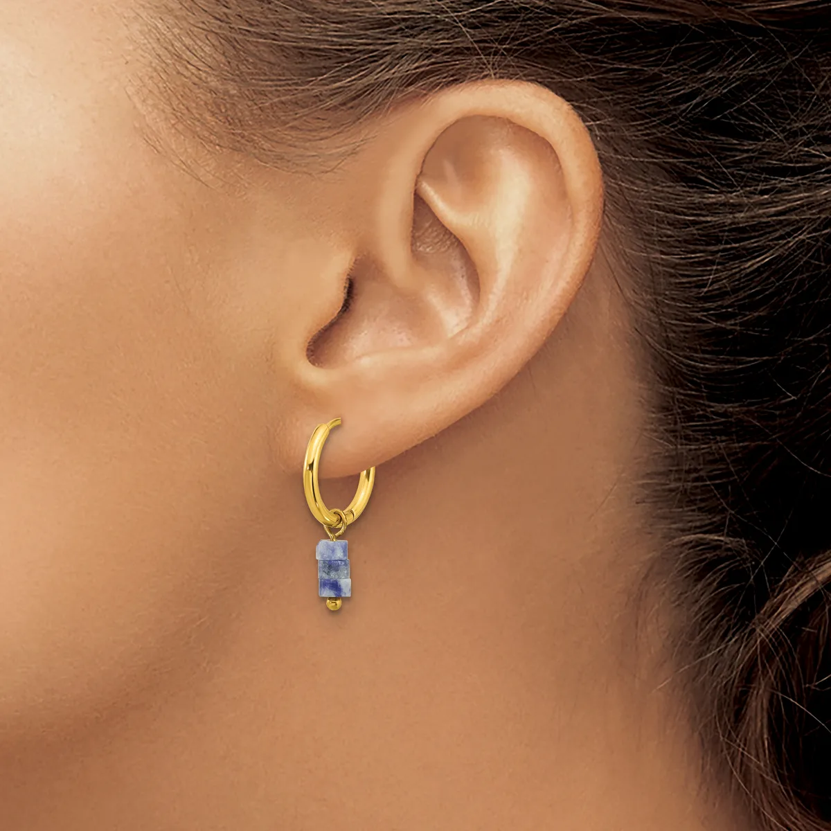 A gold hoop earring with a small rectangular blue stone pendant hangs from a woman's earlobe.