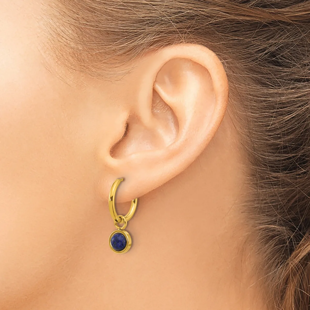 A person wearing a gold hoop earring with a small round blue stone pendant hanging from it.