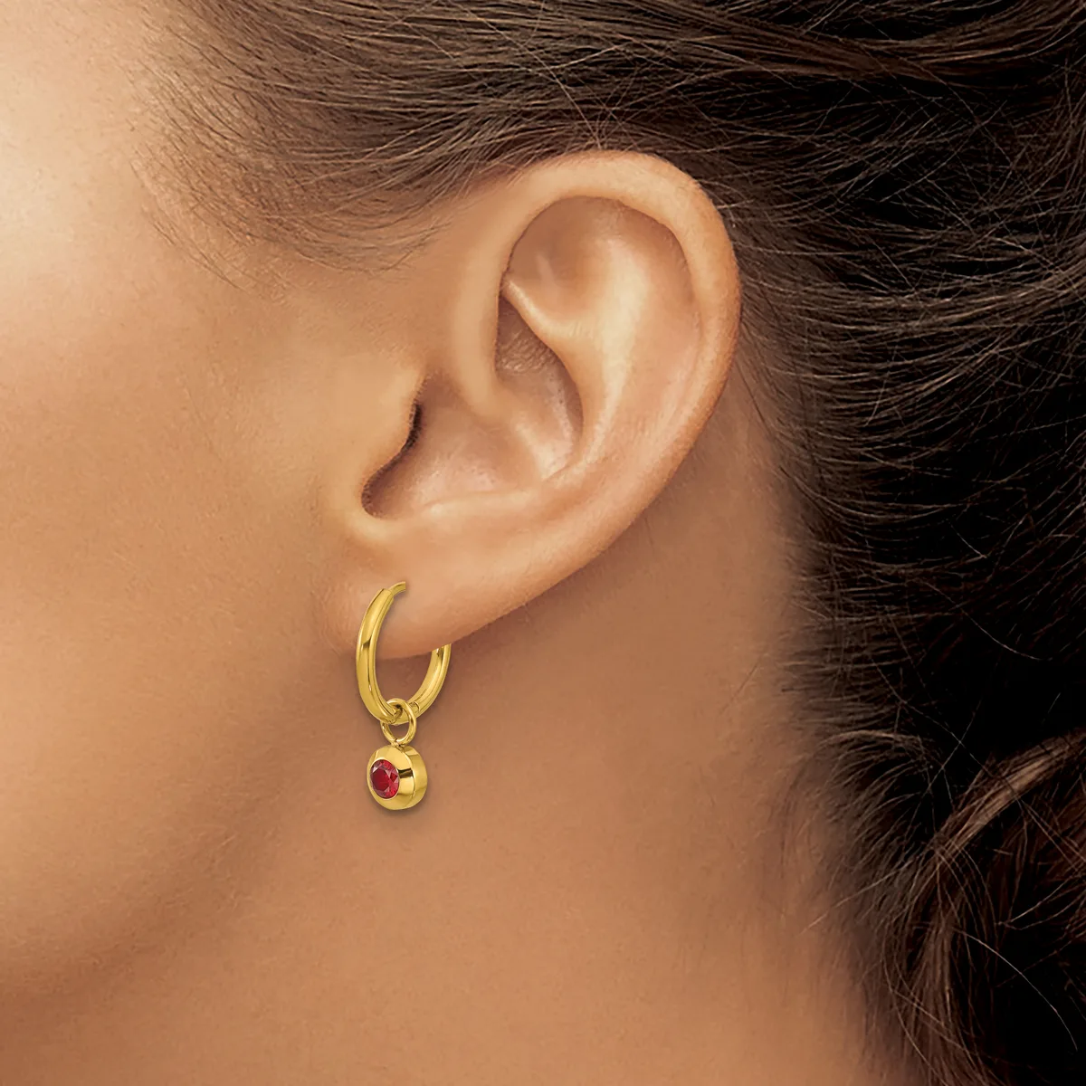 A person wearing a gold hoop earring with a small circular pendant featuring a red stone hangs from their earlobe.