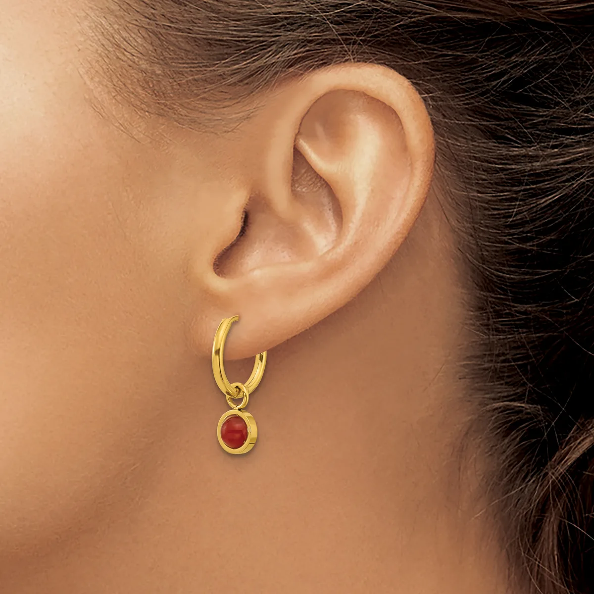 A woman wearing a gold hoop earring with a small red oval gemstone pendant hanging from it.