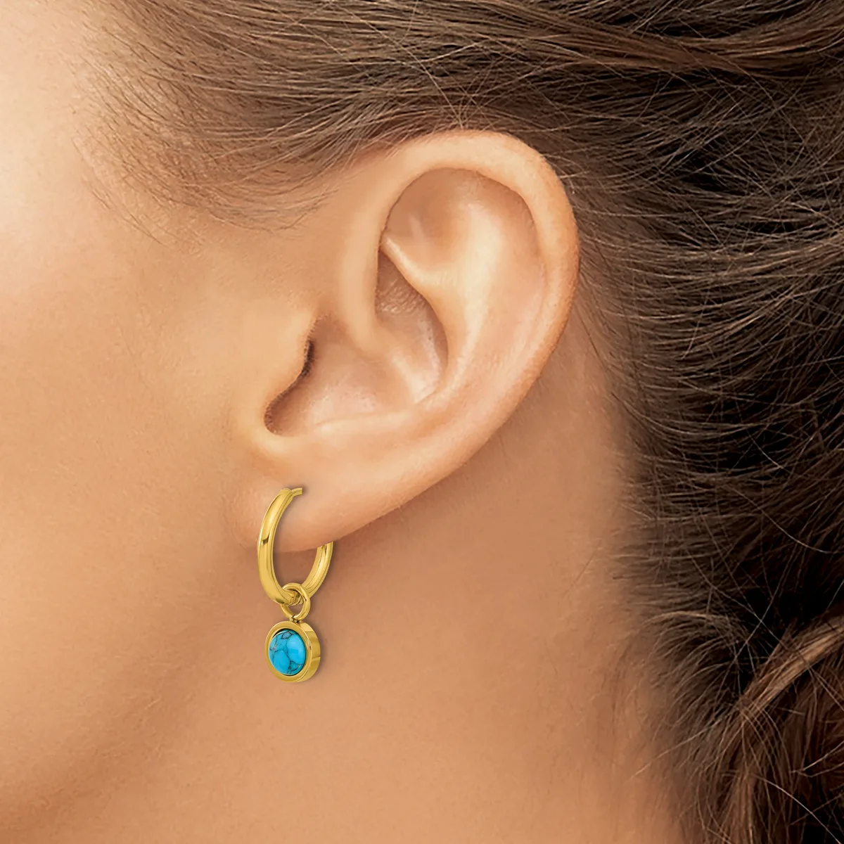 A person wearing a gold hoop earring with a small turquoise oval stone pendant hanging from it.