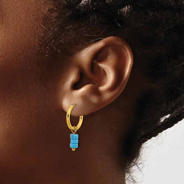 A person wearing a gold-toned hoop earring with a small turquoise blue rectangular bead charm hanging from it.