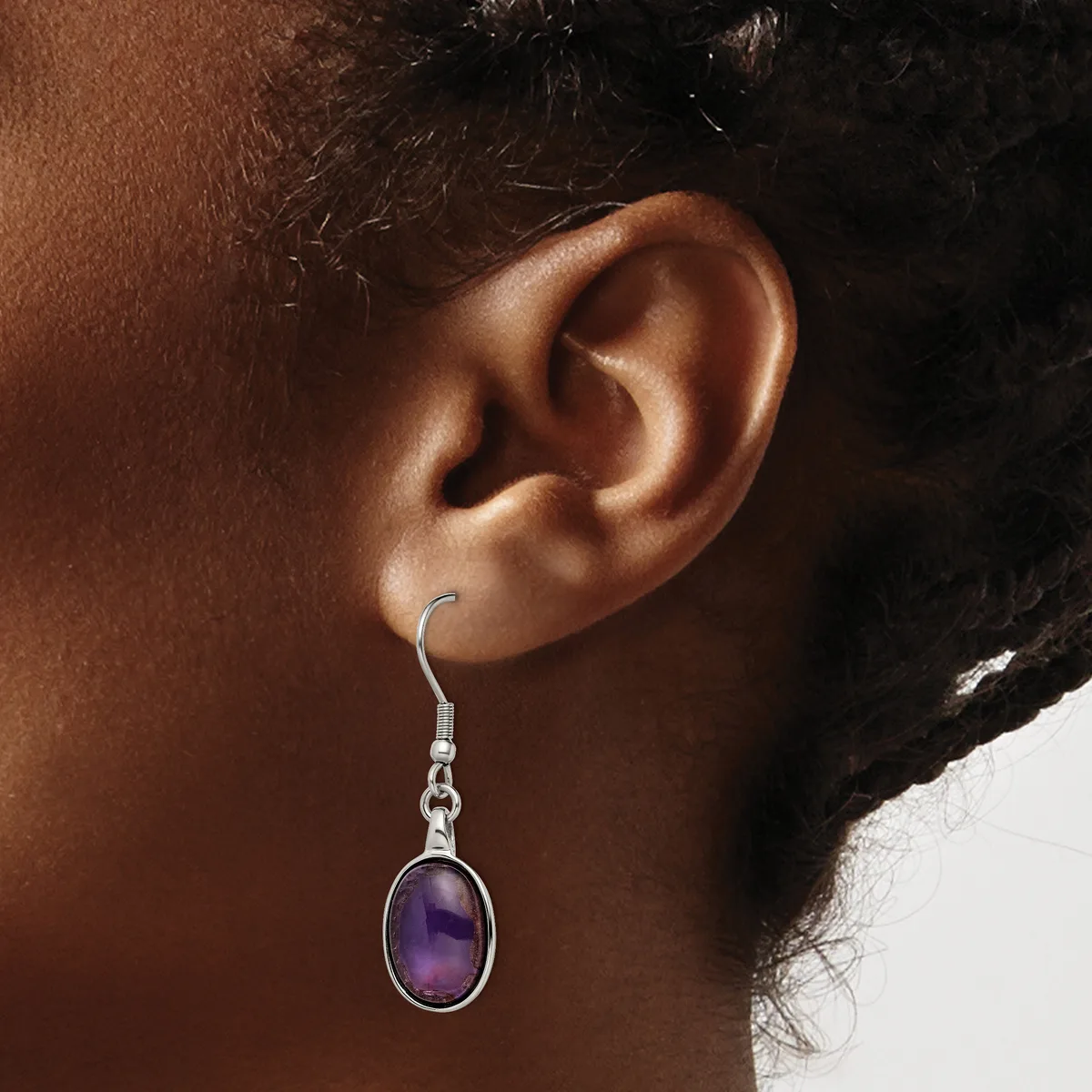 A person wears a silver dangle earring with an oval purple stone set in a bezel, suspended from a hook fastening.
