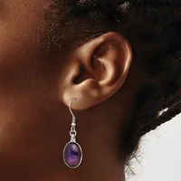 A person wears a silver dangle earring with an oval purple stone set in a bezel, suspended from a hook fastening.
