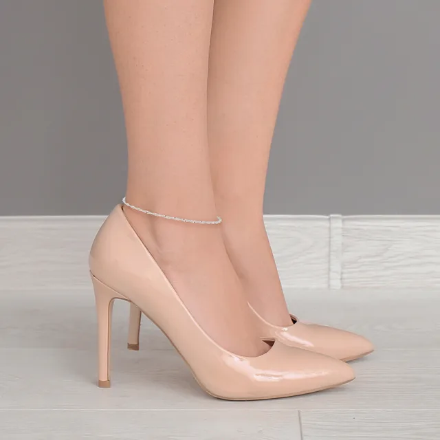 A person wearing nude patent leather high heels and a delicate beaded ankle bracelet stands against a gray and white background.