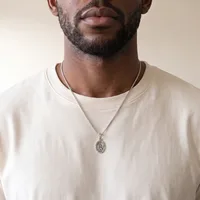 A man wearing a cream-colored t-shirt and a silver chain necklace with an oval pendant featuring an engraved design.