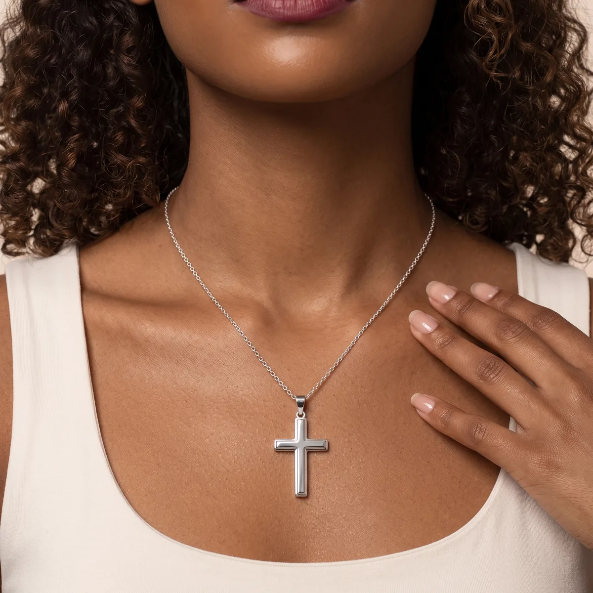 A woman wearing a white tank top displays a silver cross pendant on a delicate chain around her neck.
