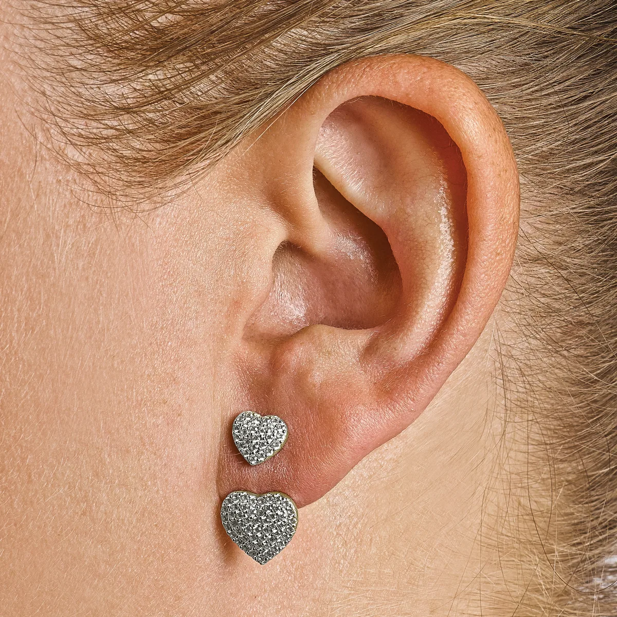 A person wearing two heart-shaped stud earrings with glittering silver surfaces in the lower and upper piercing holes of their right earlobe.