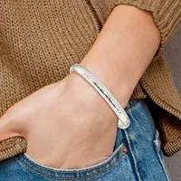 A person wearing a tan knit sweater and blue jeans displays a polished silver bangle bracelet with an engraved linear pattern on their wrist.