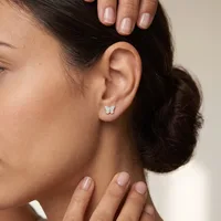 A woman wearing a small butterfly stud earring in her earlobe, with dark hair styled in a bun and natural-looking manicured nails.