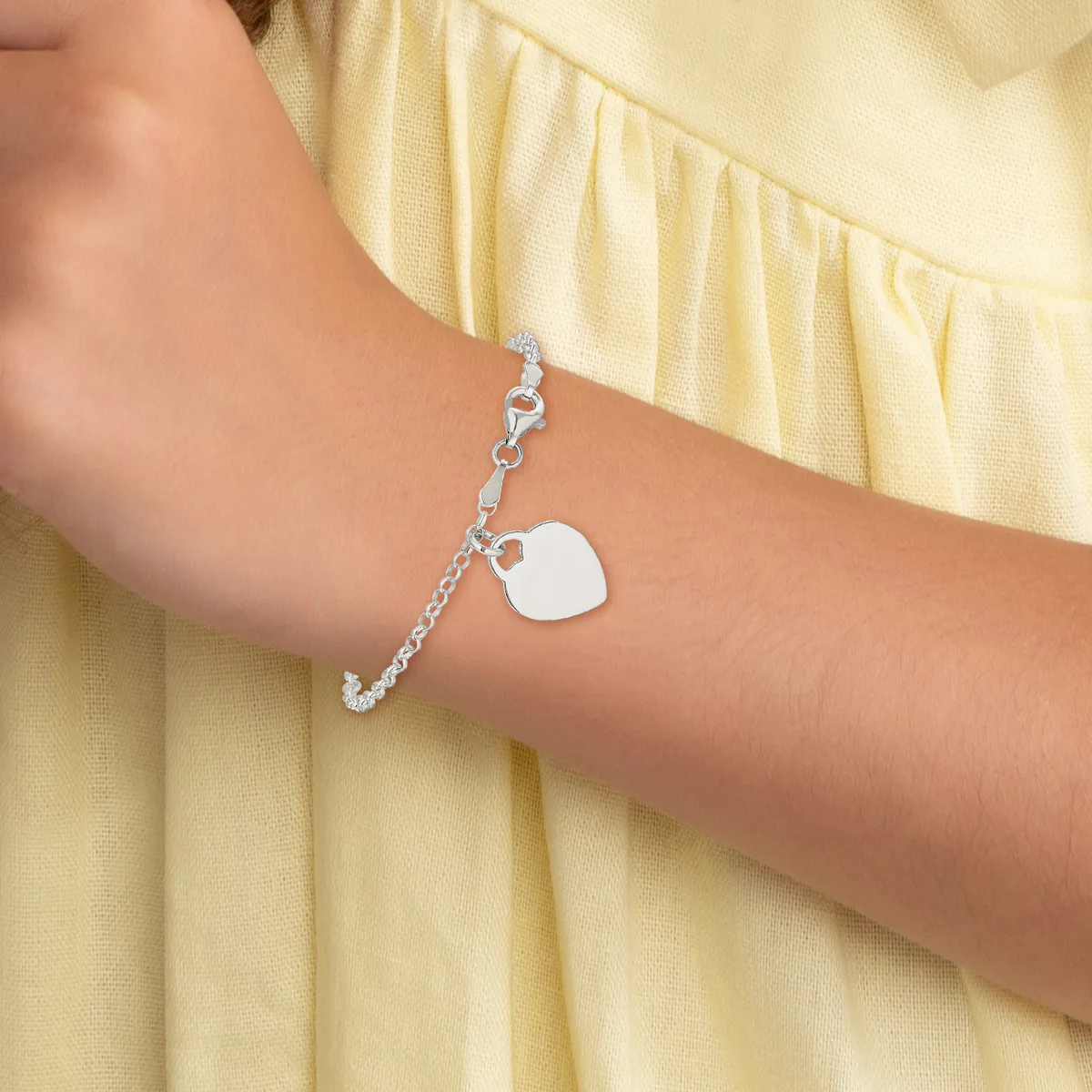 A silver chain bracelet with a blank square charm hangs from the wrist of a person wearing a pale yellow ruffled sleeve.