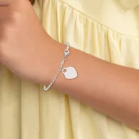 A silver chain bracelet with a blank square charm hangs from the wrist of a person wearing a pale yellow ruffled sleeve.