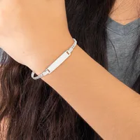A woman wearing a gray v-neck shirt displays a silver-tone bracelet with a rectangular nameplate and decorative beaded chain links on her wrist.
