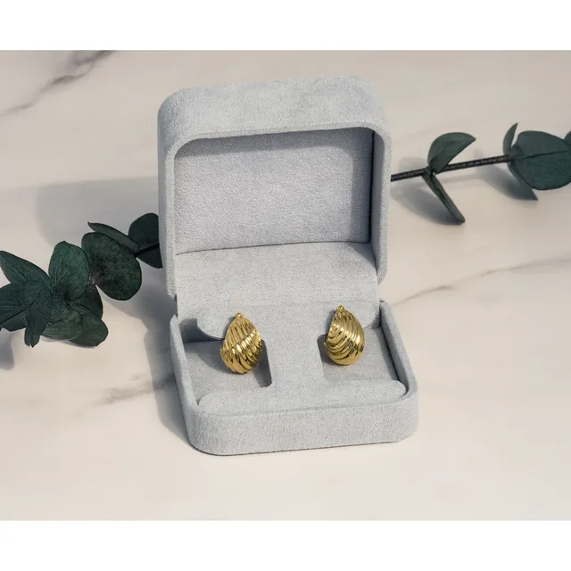 A pair of gold teardrop-shaped stud earrings displayed in an open light gray jewelry box, surrounded by green eucalyptus branches on a marble surface.