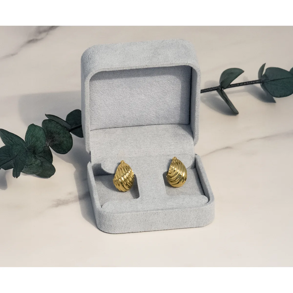 A pair of gold teardrop-shaped stud earrings displayed in an open light gray jewelry box, surrounded by green eucalyptus branches on a marble surface.