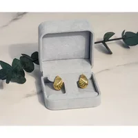 A pair of gold teardrop-shaped stud earrings displayed in an open light gray jewelry box, surrounded by green eucalyptus branches on a marble surface.