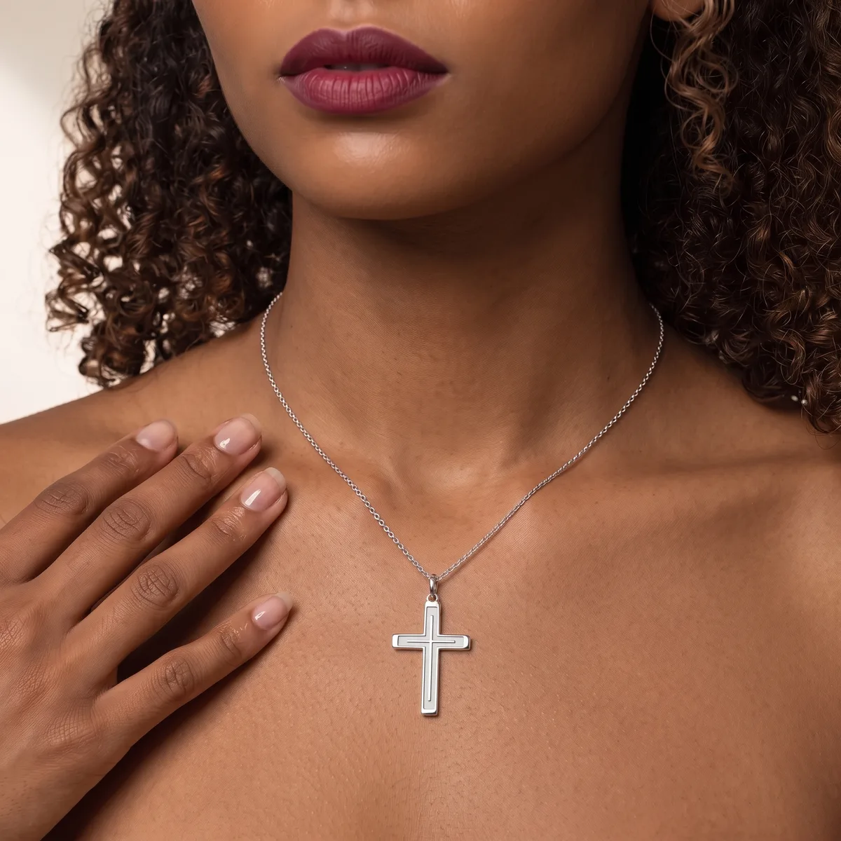Woman wearing a silver cross pendant necklace on a chain, with curled dark hair and mauve lipstick, hand placed near chest.