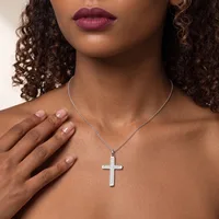 Woman wearing a silver cross pendant necklace on a chain, with curled dark hair and mauve lipstick, hand placed near chest.