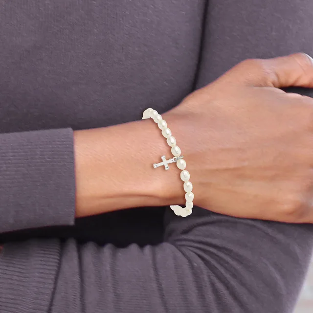 A woman wearing a gray long-sleeved shirt displays a beaded bracelet on her wrist with white pearl-like beads and a small silver cross charm.