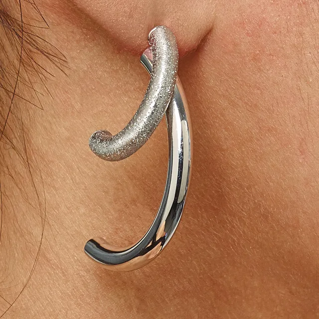 A curved silver earring with a textured top and polished bottom portion hangs from an earlobe against bare skin.