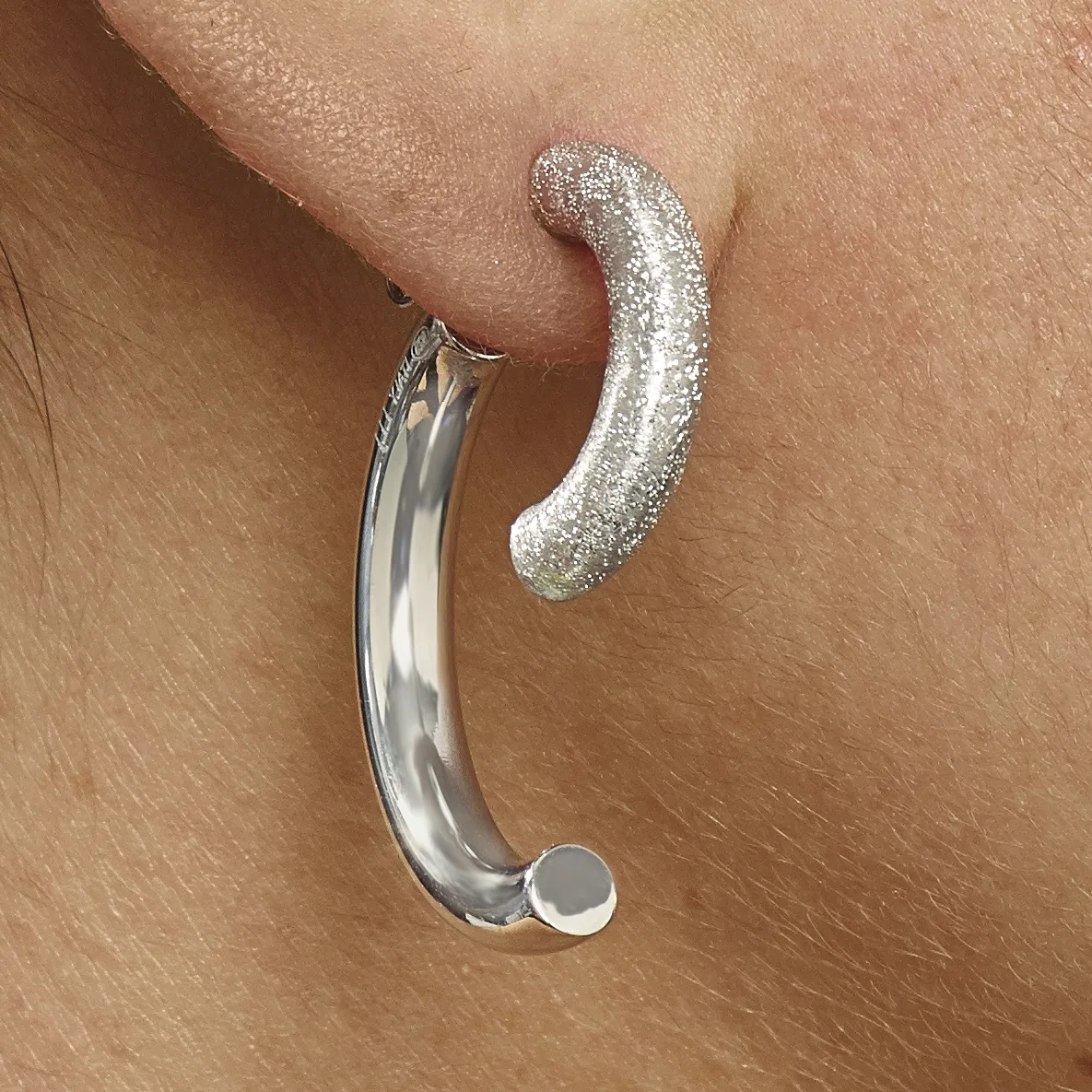 Two curved silver earrings worn on a pierced ear, one plain polished and one textured with glitter finish.