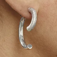 Two curved silver earrings worn on a pierced ear, one plain polished and one textured with glitter finish.