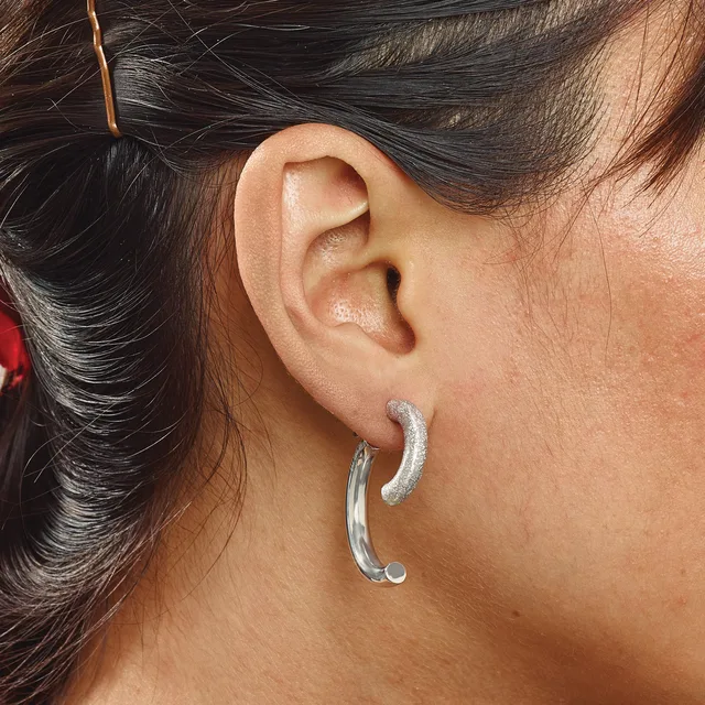 A woman wearing curved silver hoop earrings with a textured finish, shown from the side with her dark hair pulled back.