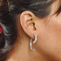 A woman wearing curved silver hoop earrings with a textured finish, shown from the side with her dark hair pulled back.