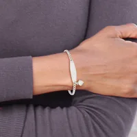 A person wearing a gray long-sleeved top displays a silver-toned bangle bracelet with a white rectangular pendant and small charm on their wrist.