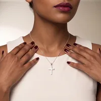 A woman wearing a white sleeveless top and a silver chain necklace with a cross pendant displays her manicured nails with dark burgundy polish.