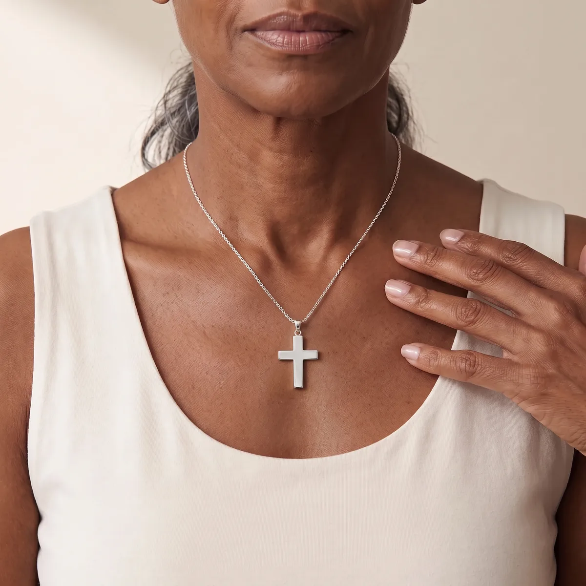 A person wearing a white sleeveless tank top displays a silver cross pendant on a delicate chain, resting at the neckline.