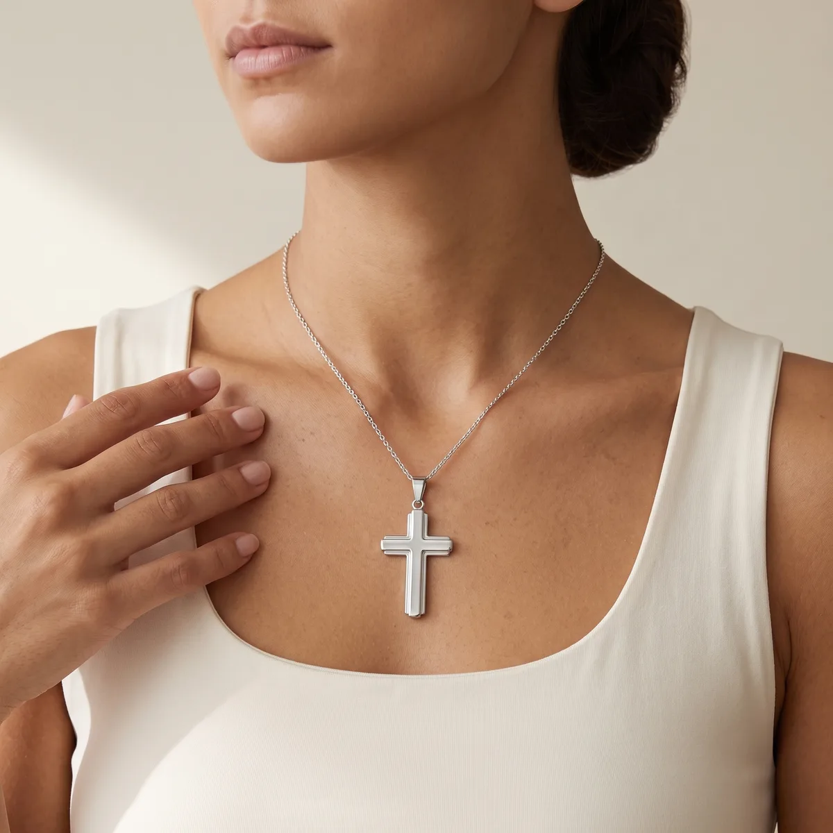 A woman wearing a white sleeveless top displays a silver cross pendant necklace hanging at her chest.