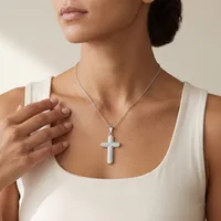 A woman wearing a white sleeveless top displays a silver cross pendant necklace hanging at her chest.