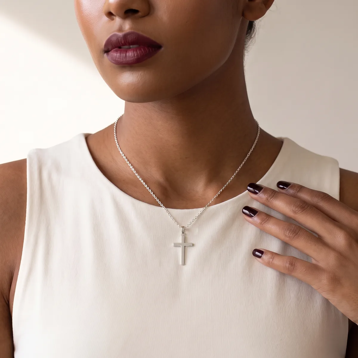 A woman wearing a cream sleeveless top and silver chain necklace with a cross pendant, displaying manicured nails with dark polish and burgundy lipstick.
