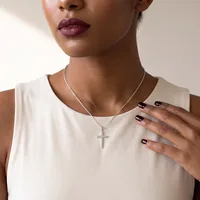 A woman wearing a cream sleeveless top and silver chain necklace with a cross pendant, displaying manicured nails with dark polish and burgundy lipstick.