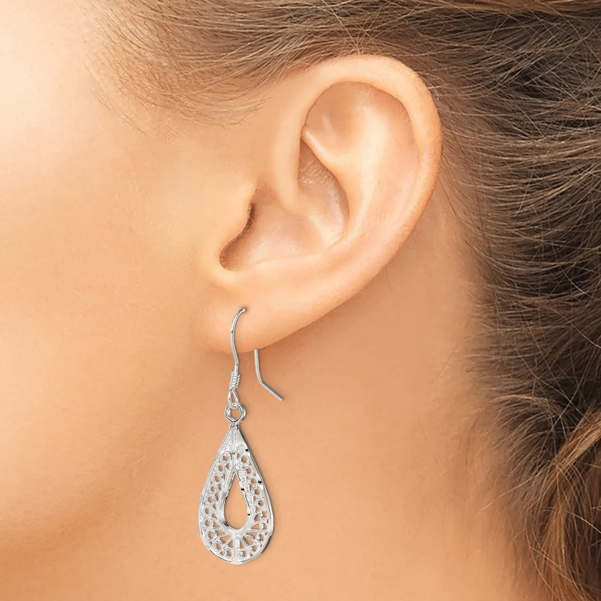 A woman wearing a teardrop-shaped silver dangle earring with a openwork filigree pattern and rhinestone embellishments on a fish hook back.
