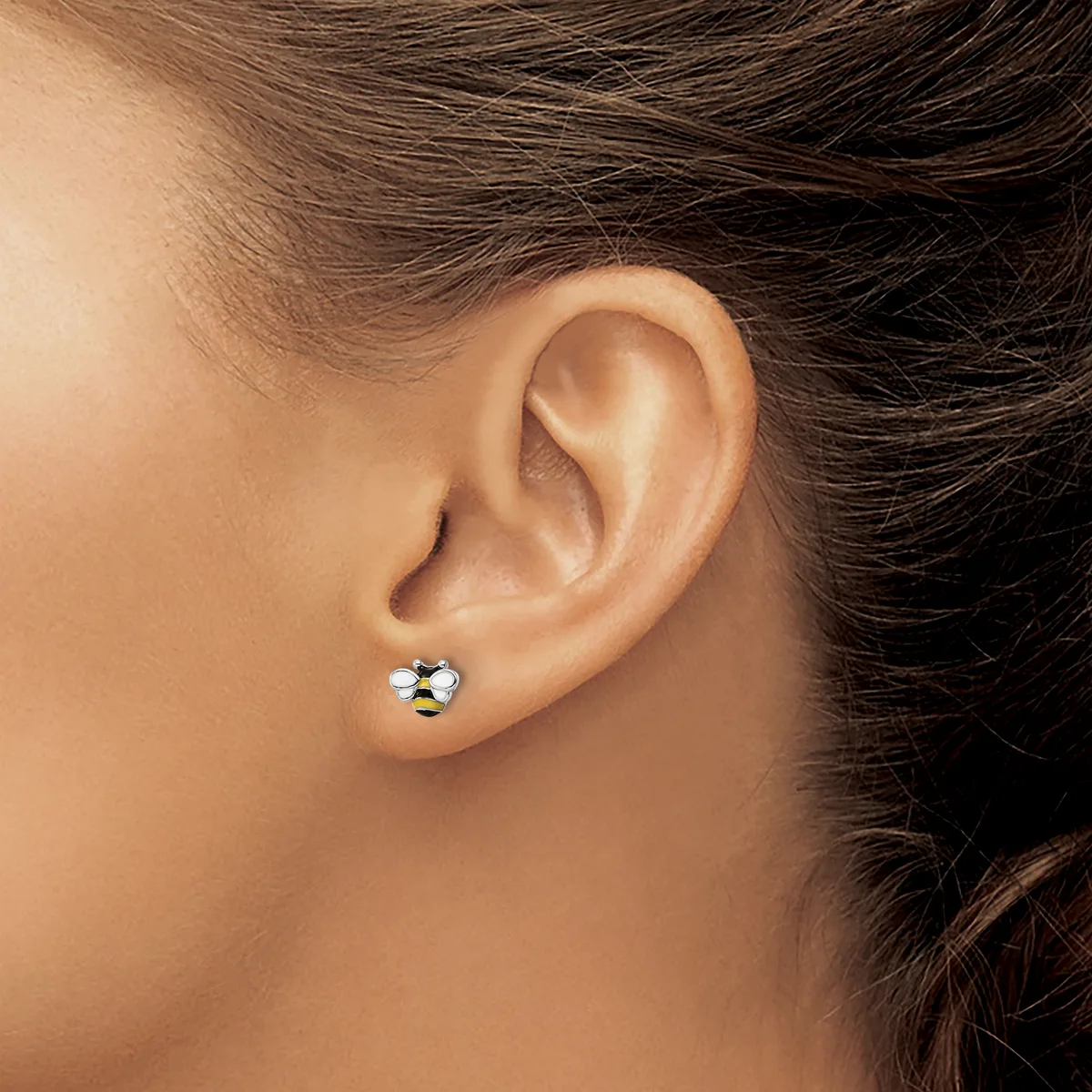 A person wearing a small stud earring with a bee design in gold and white enamel in their pierced earlobe.