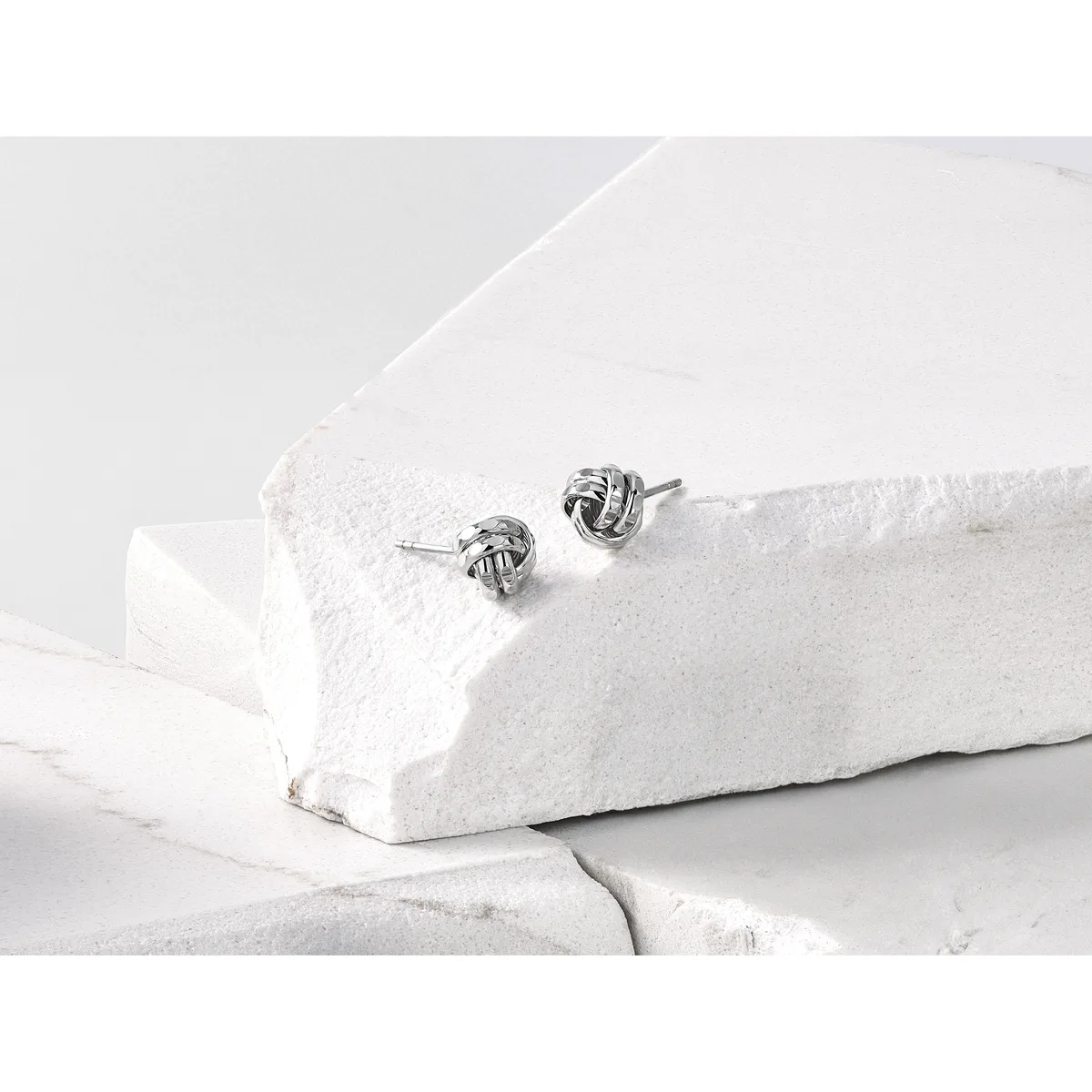 Pair of silver knot stud earrings displayed on white textured stone blocks.