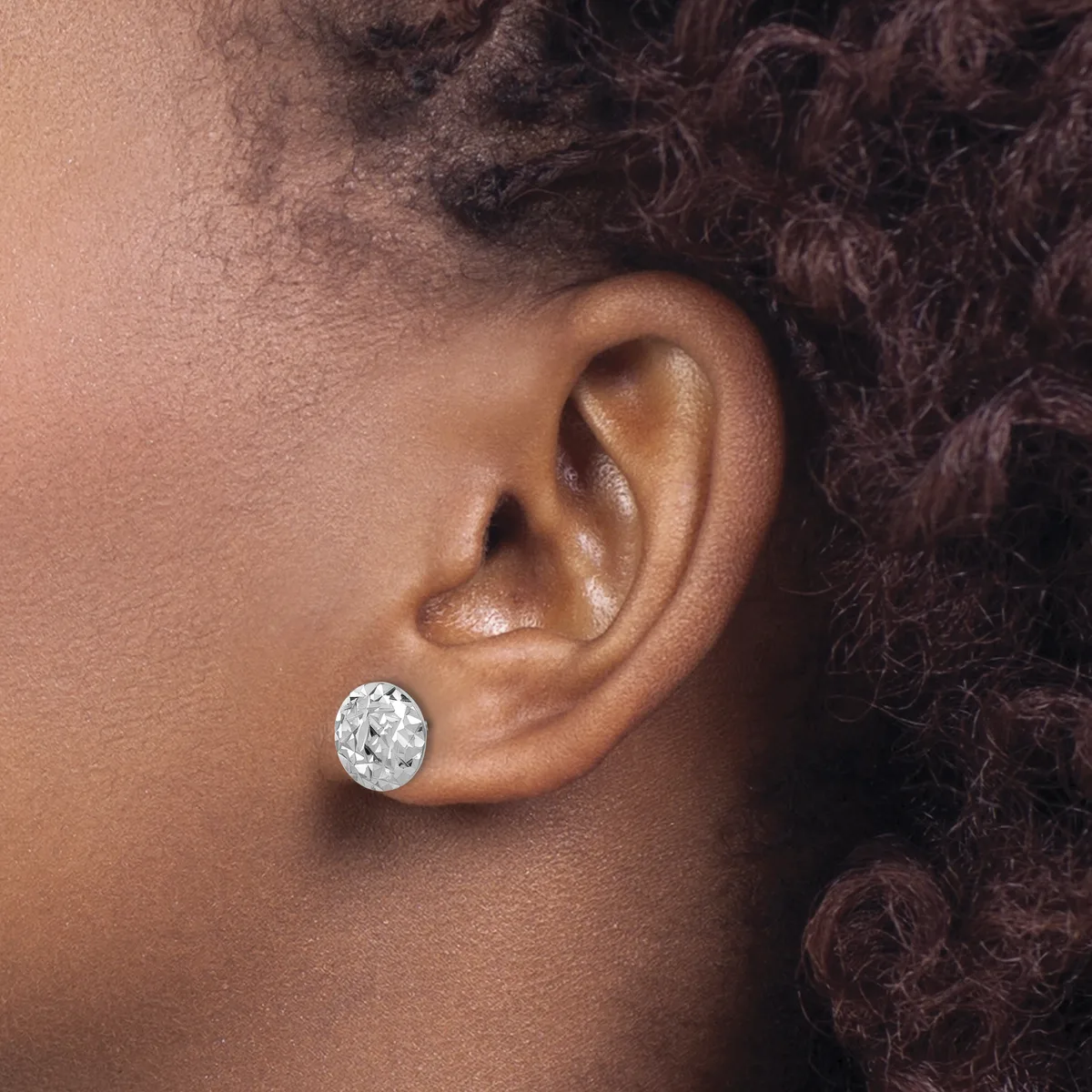A person wearing a single round diamond stud earring in their pierced earlobe, shown in profile view against their shoulder and dark curly hair.
