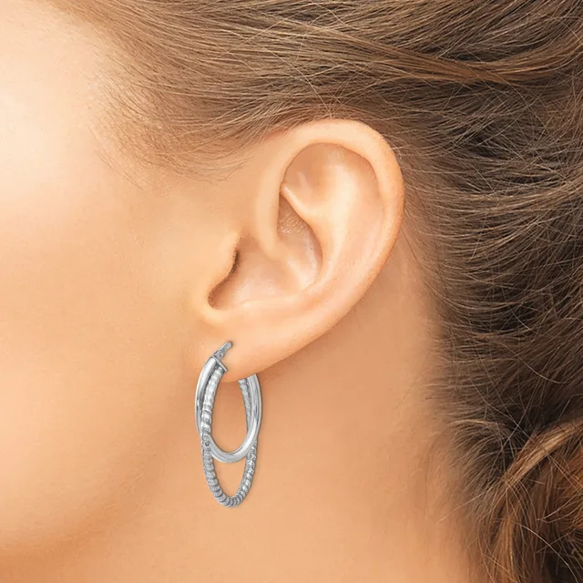 A woman wears a pair of textured silver-toned hoop earrings with a double-loop design, shown from the side view of her ear.