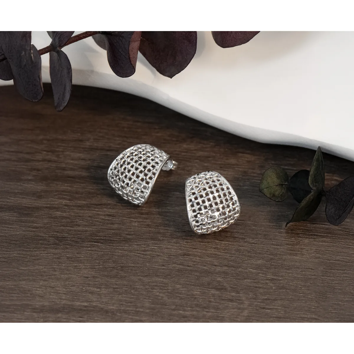 Two silver mesh dome-shaped earrings displayed on a dark wooden surface next to a white bowl and dark green eucalyptus leaves.