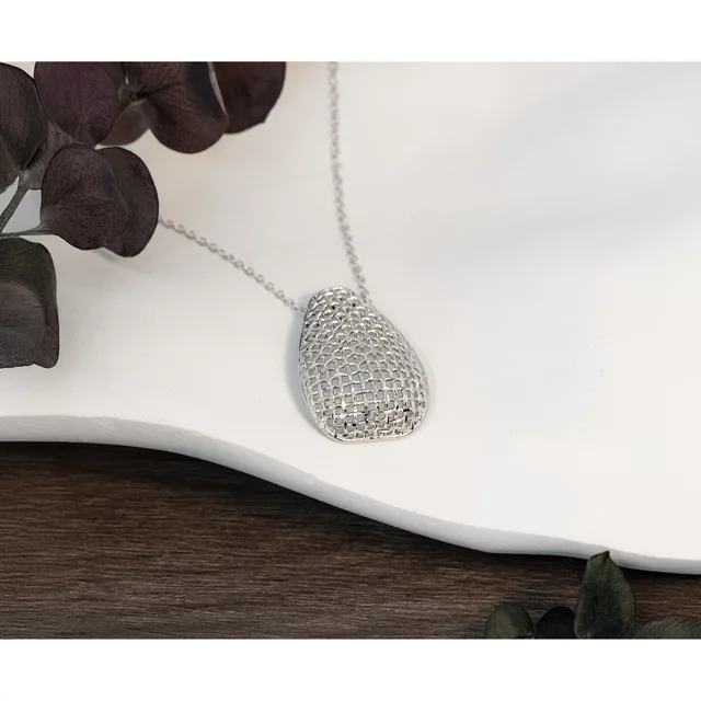 Silver teardrop pendant with textured mesh pattern on a delicate chain, displayed on white ceramic dish with dark foliage beside it.