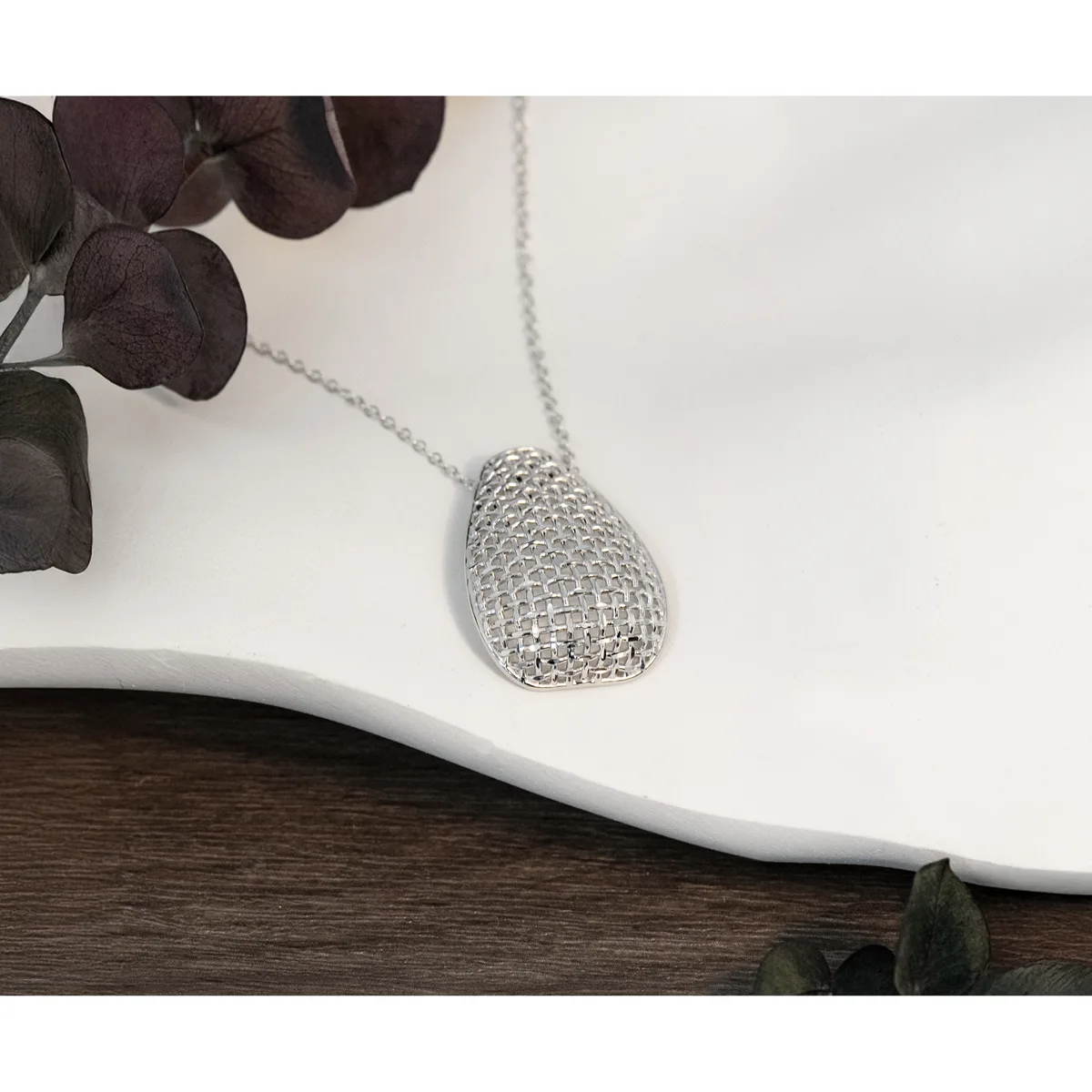 Silver teardrop pendant with textured mesh pattern on a delicate chain, displayed on white ceramic dish with dark foliage beside it.