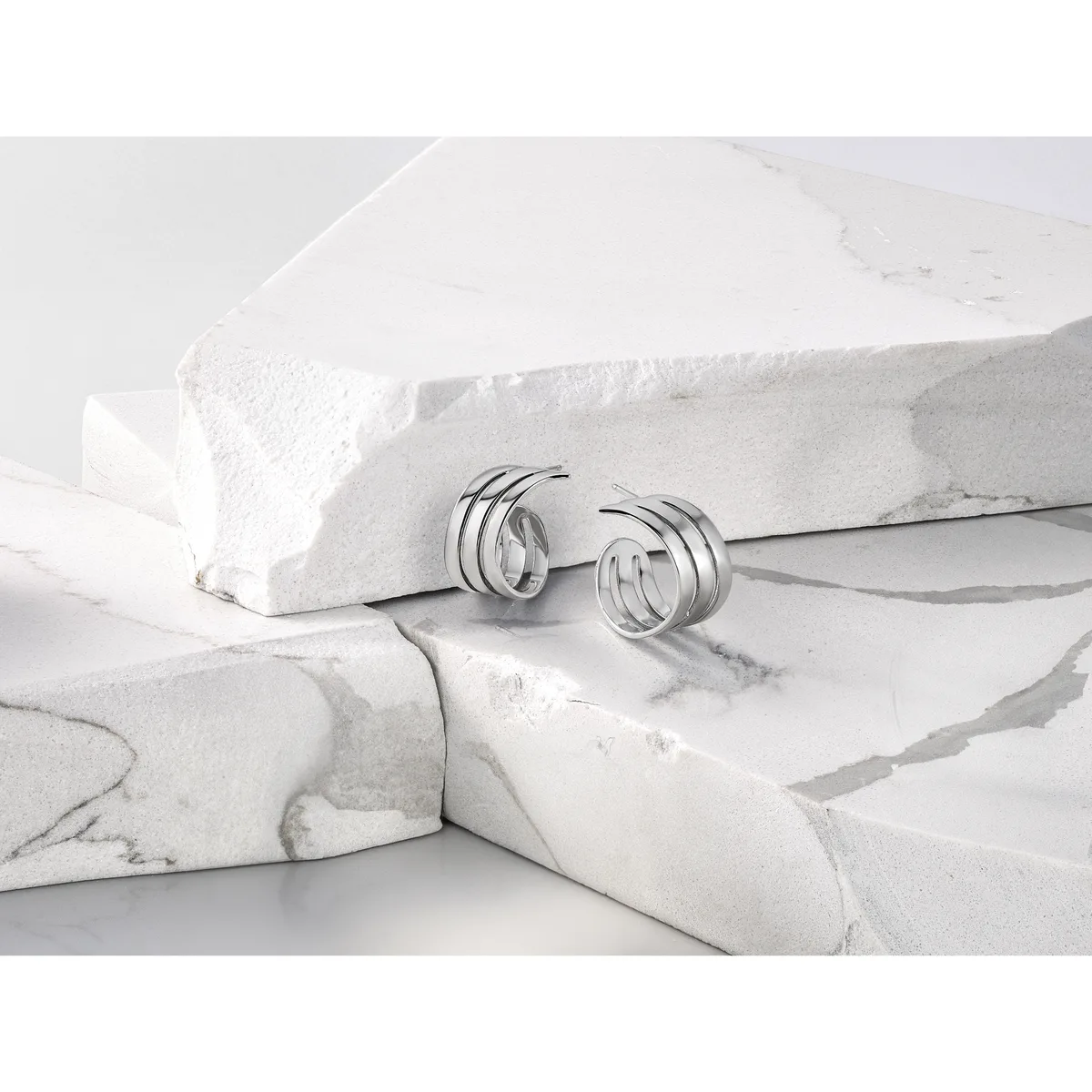 Silver hoop earrings with triple-band design displayed on white marble surfaces and draped fabric.