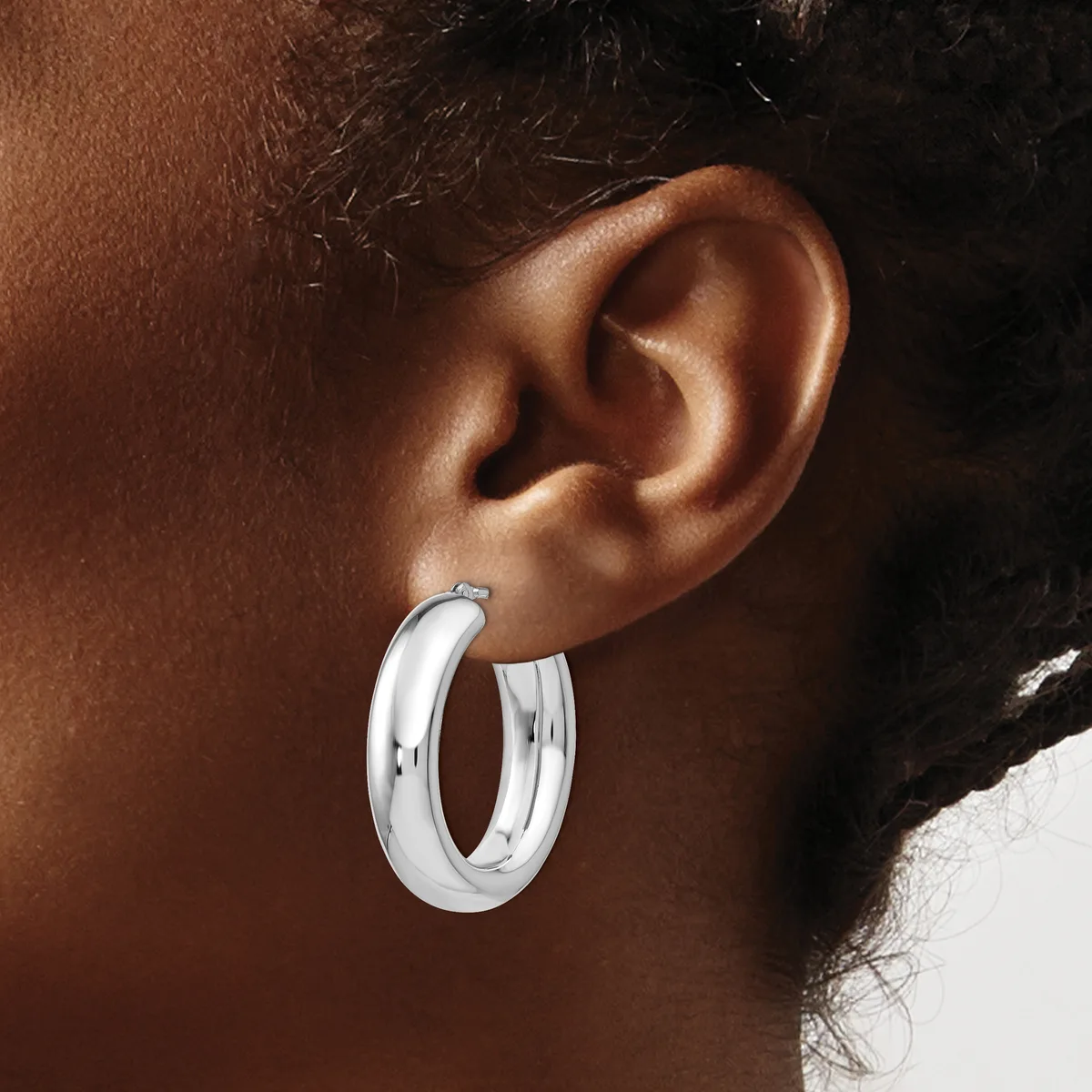 A person wearing a pair of polished silver hoop earrings with a rounded, chunky profile, shown from the side view against their ear.